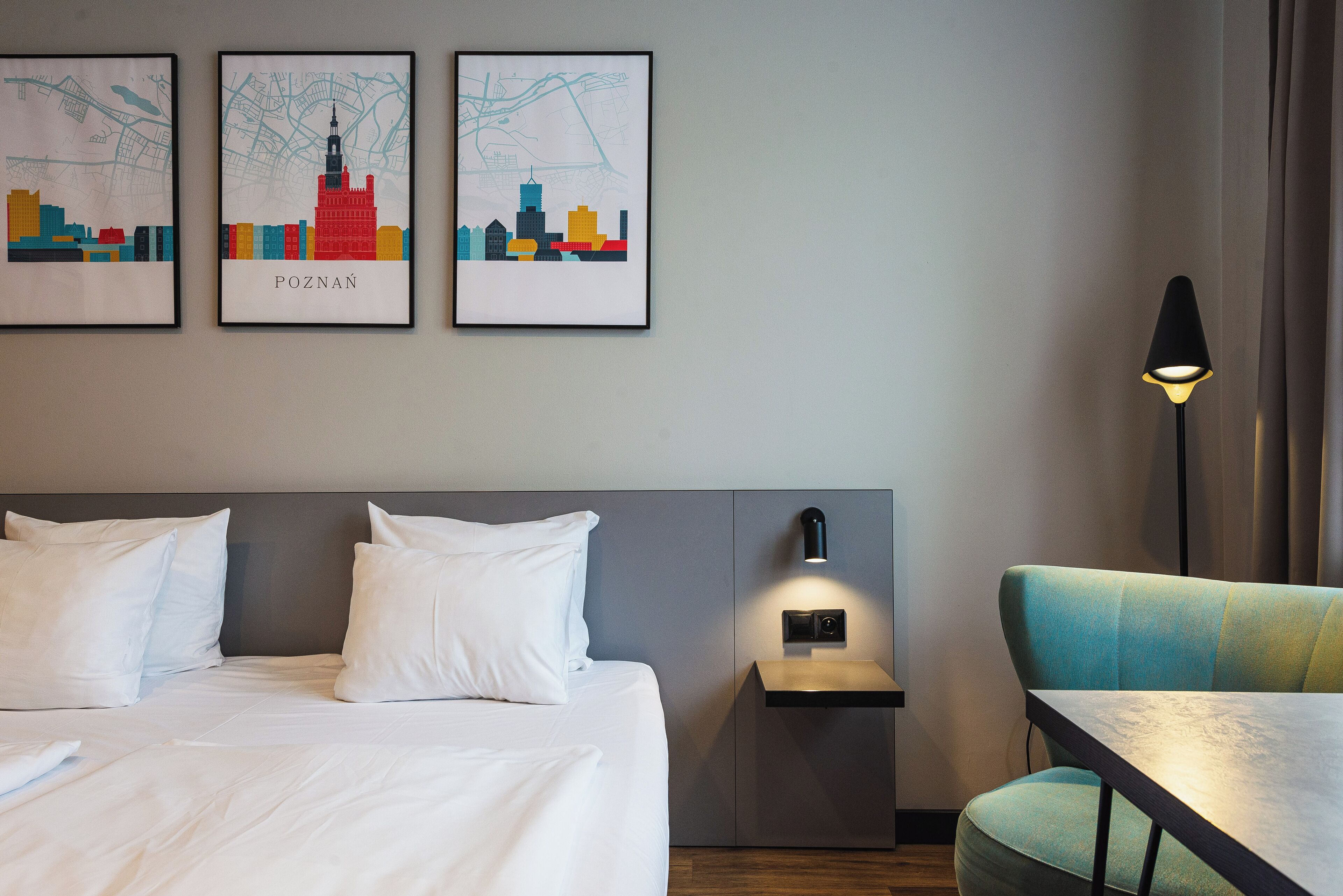 A hotel room with a bed, pillows, a nightstand, a lamp, and two framed cityscape pictures on the wall.
