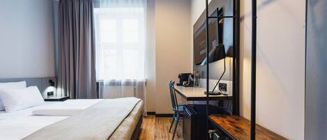 A hotel room with a bed, a desk, a chair, a small kitchenette, and a window with curtains.