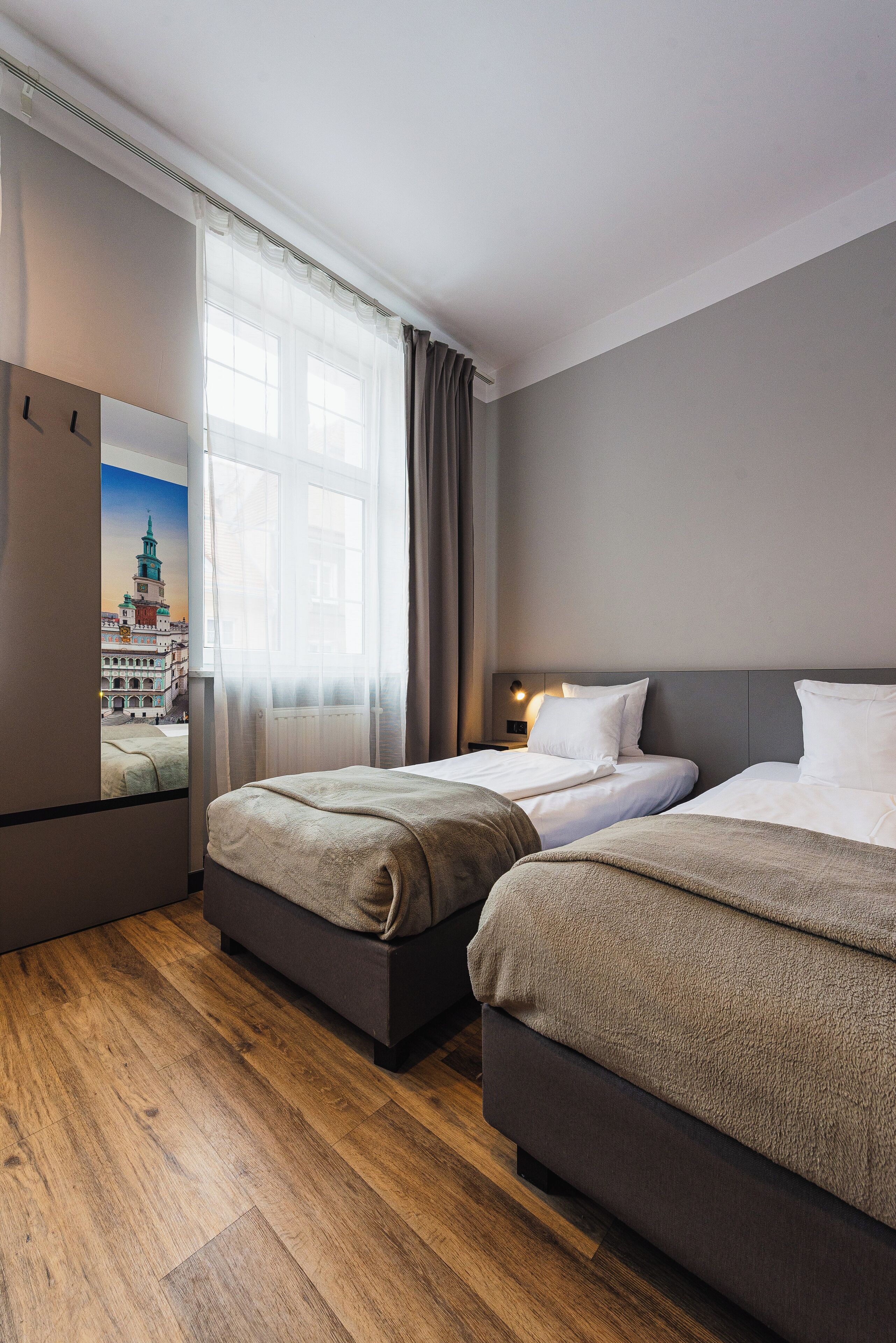Une chambre d’hôtel avec deux lits, un plancher en bois, une grande fenêtre avec des rideaux et un téléviseur fixé au mur qui diffuse une vue sur la ville.