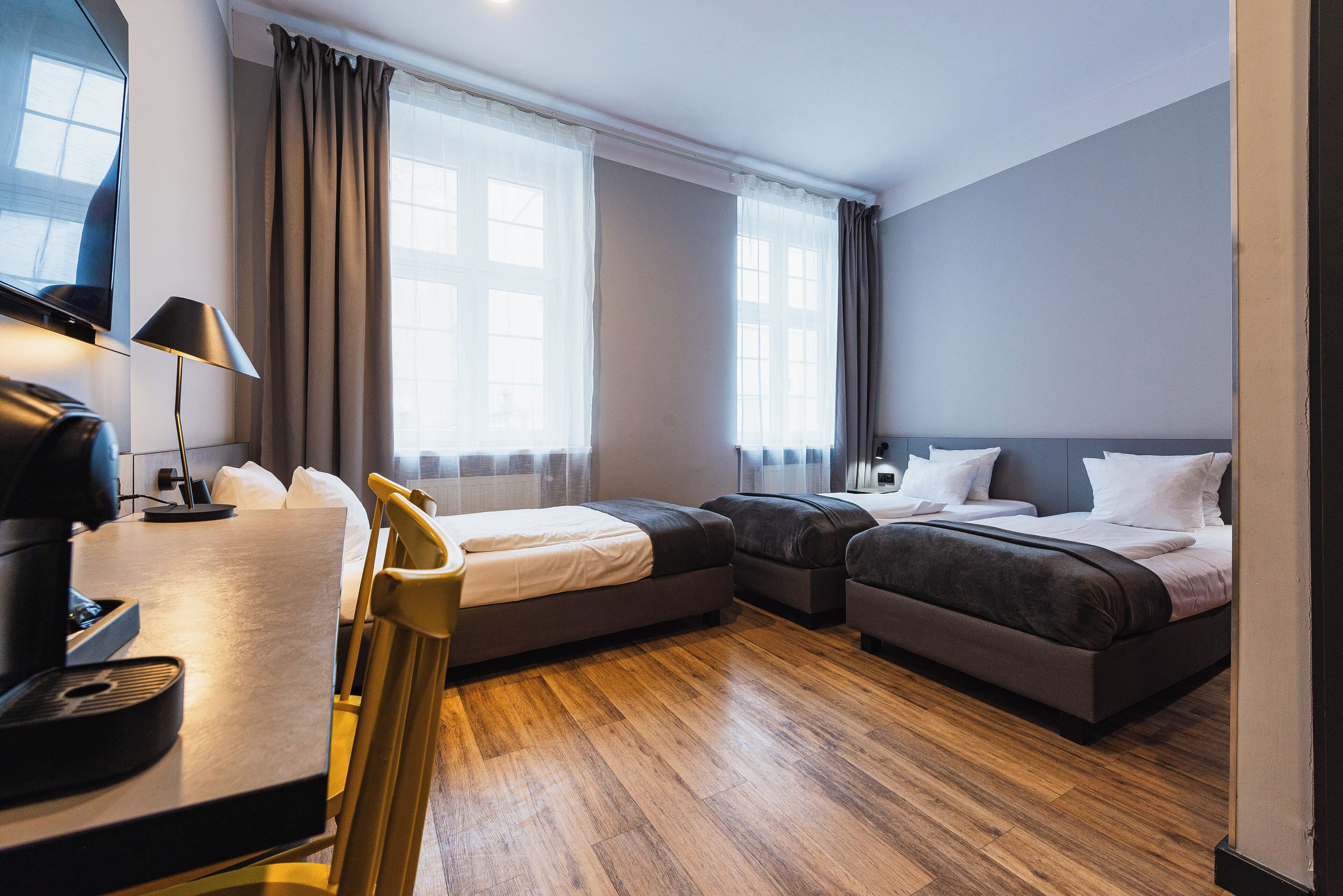 A hotel room with a desk, two beds, a TV, and large windows with curtains.