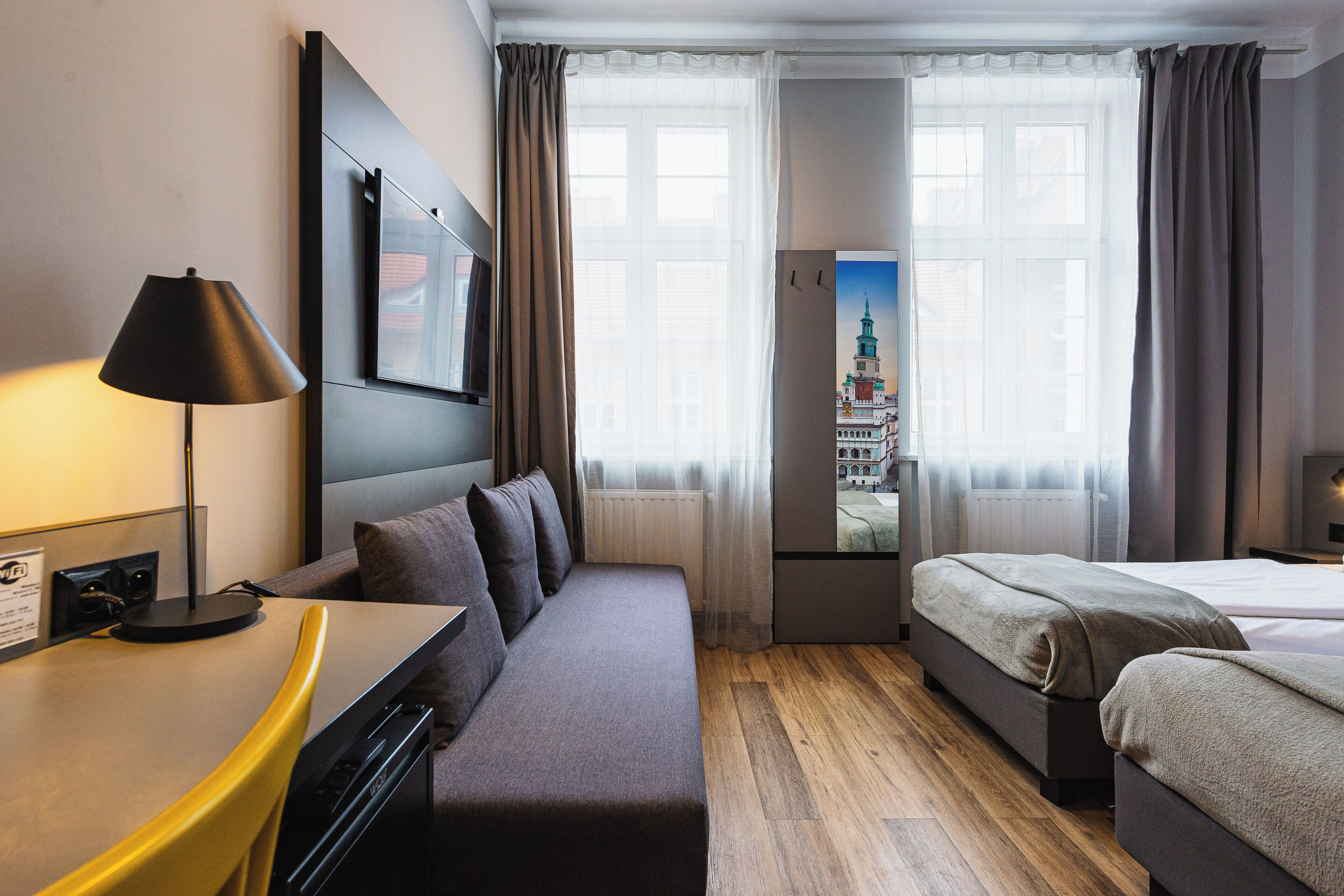 A hotel room with a sofa bed, a desk with a lamp, a bed, and a view of a cityscape through the window.