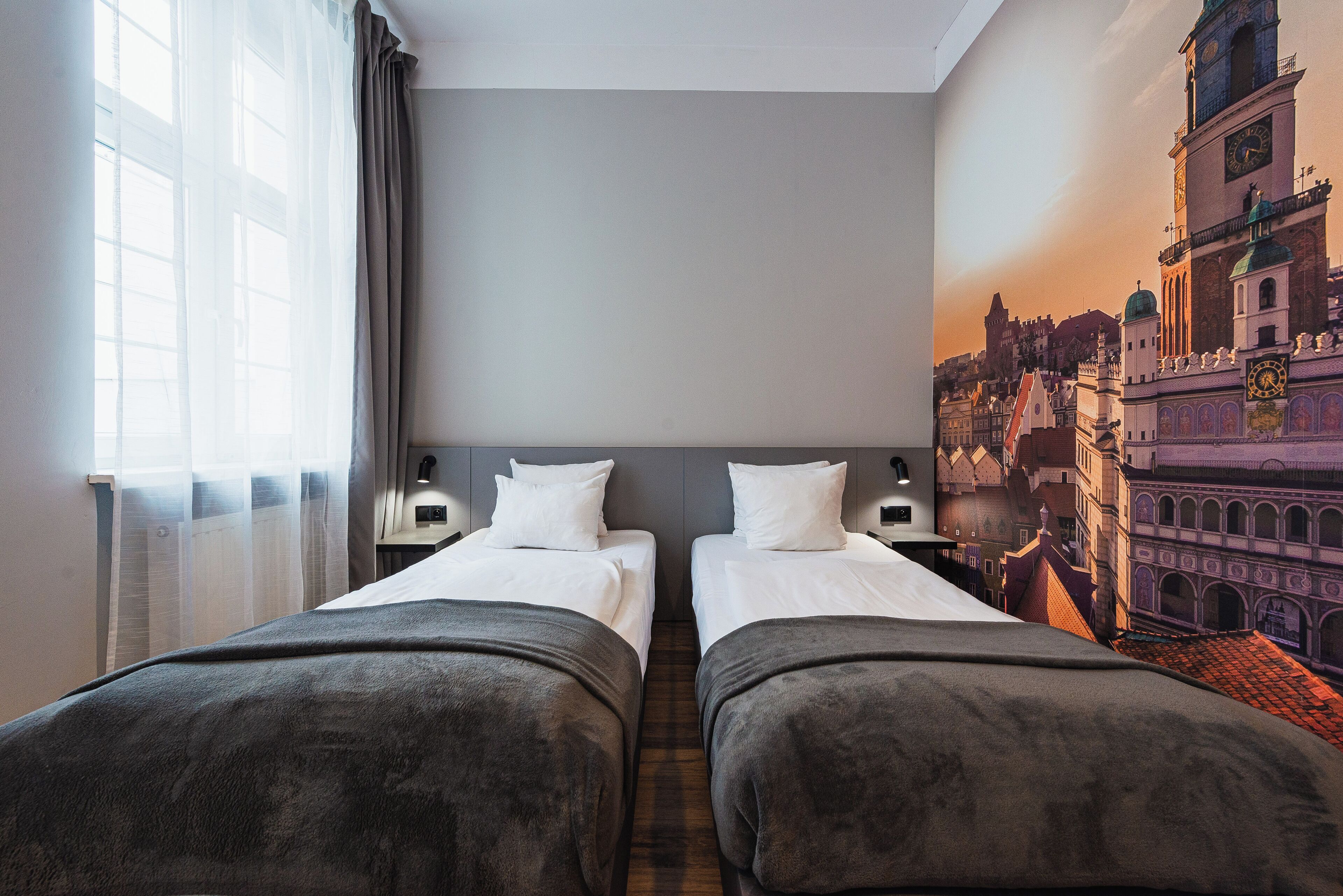 Habitación de hotel con dos camas individuales, un ventanal con cortinas translúcidas y un mural con vista a la ciudad en la pared.