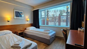 A hotel room with two beds, a wooden headboard, a window with a view of trees, and a table with a chair. - Hotel Austur (Fjardabyggd)