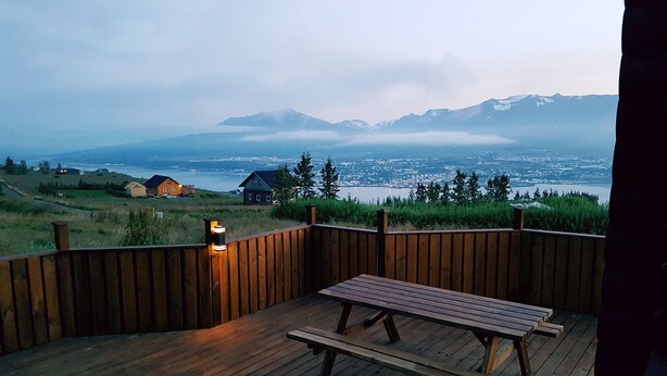 A wooden deck with a picnic table and a light fixture, overlooking a body of water and distant mountains. - Akureyri Log Cabin (Akureyri)