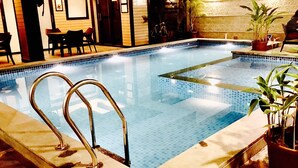 An indoor swimming pool with clear water, surrounded by a tiled deck. There are two metal pool ladders on the left side. The pool area is adjacent to a room with a wooden floor and a door. - Le Magnifique (Alto de Porvorim)