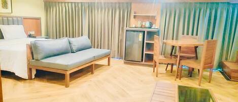 Habitación de hotel con cama, sofá, mesa de comedor con sillas, una pequeña cocina americana y un amplio ventanal con cortinas.