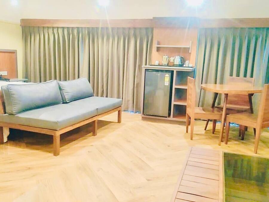 Habitación de hotel con cama, sofá, mesa de comedor con sillas, una pequeña cocina americana y un amplio ventanal con cortinas.