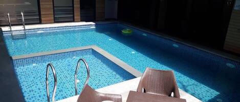 A swimming pool with two metal ladders, surrounded by brown wicker chairs and a table.
