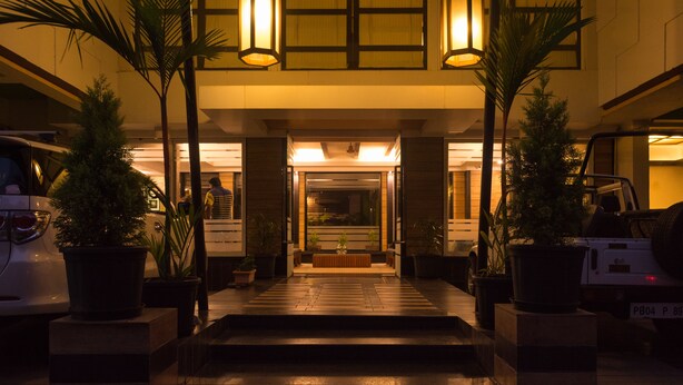 A hotel entrance with a staircase leading to a reception area, flanked by potted plants and illuminated by overhead lights. - Le Magnifique (Alto de Porvorim)