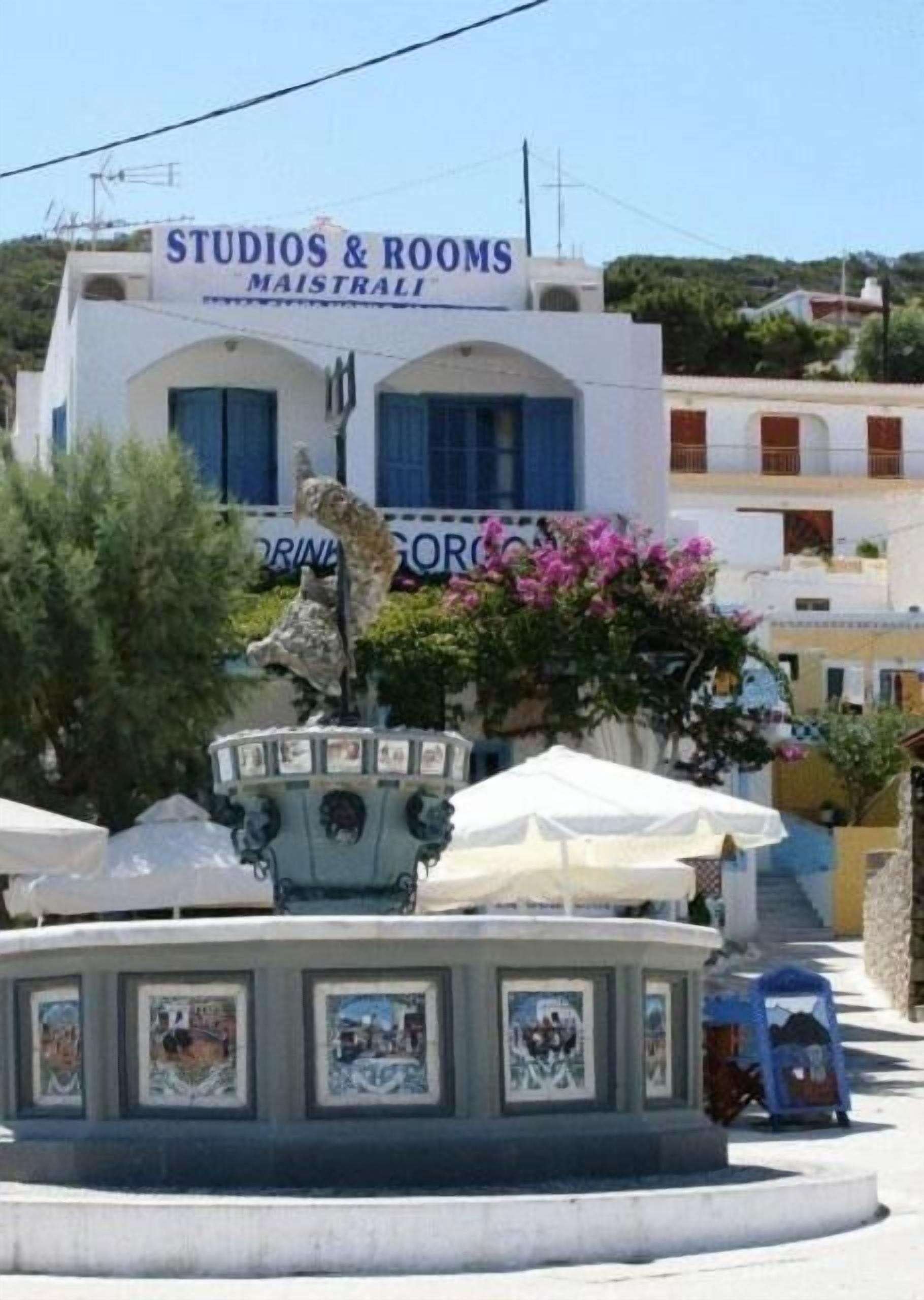 A building with a sign reading "STUDIOS & ROOMS MAISTRALI" and a fountain with a statue in front.