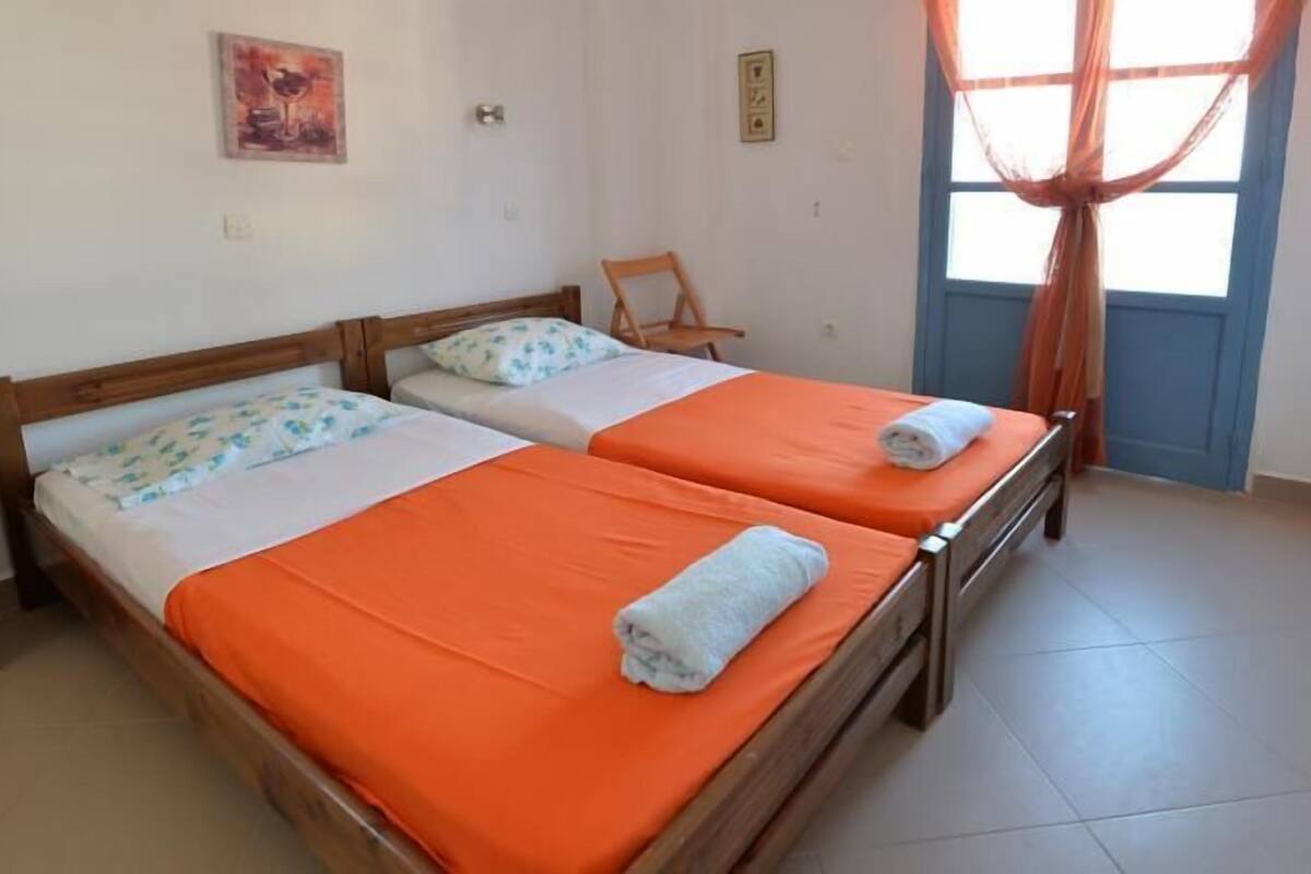A room with two wooden beds, each with orange bedding and white pillows. There is a wooden nightstand on the left, a chair in the corner, and a window with curtains.