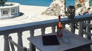Een terras aan de zee met een stenen fontein, een tafel met een fles en glazen, en uitzicht op de zee en een rotsachtig eiland.