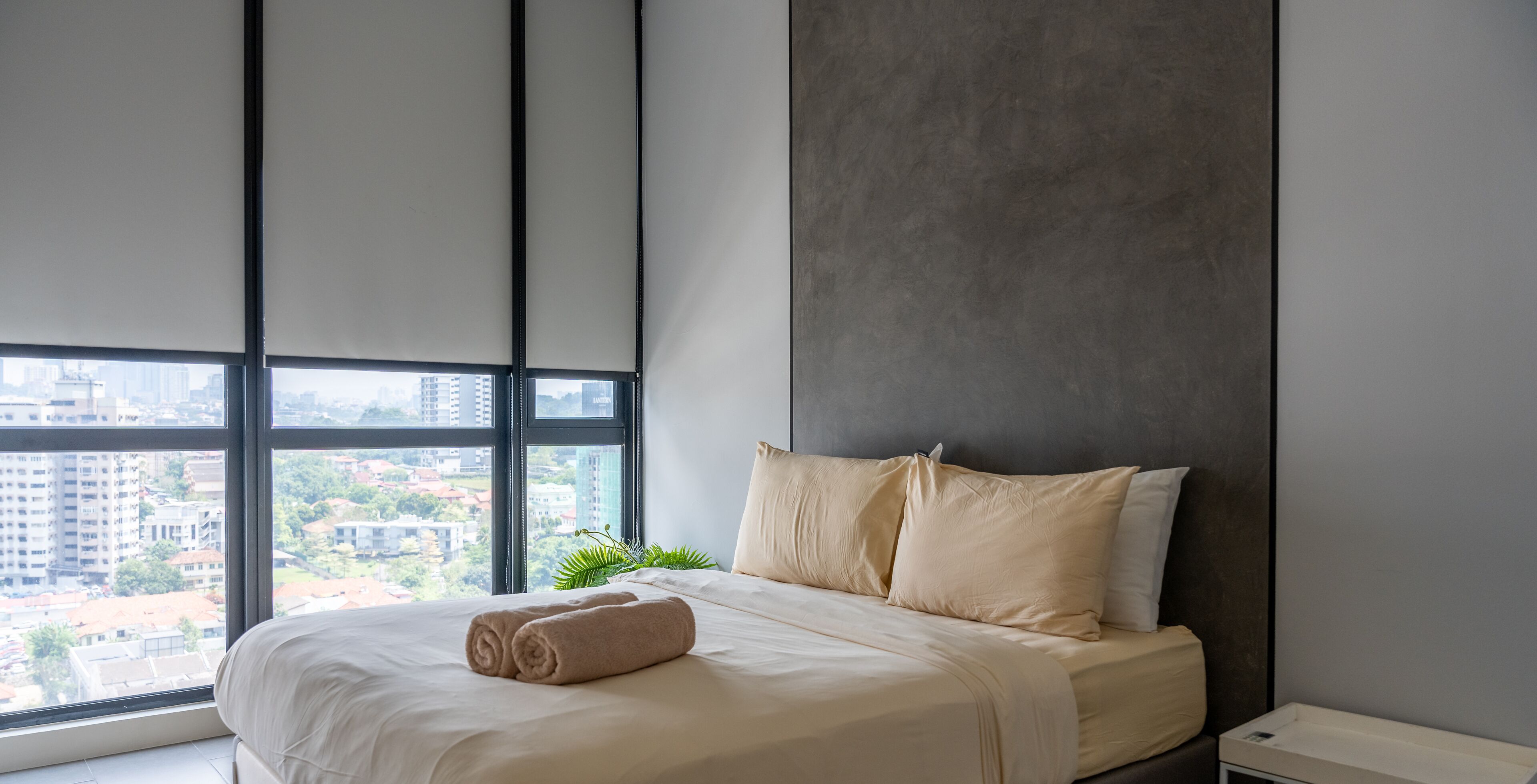 Une chambre d’hôtel moderne dotée d’une grande fenêtre donnant sur la ville, d’un lit bien fait avec du linge de lit beige et d’un design minimaliste.