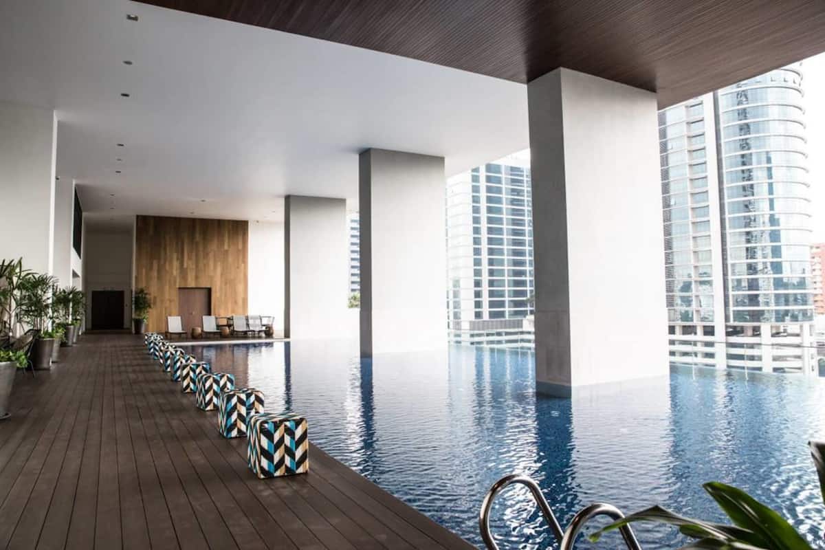 A modern indoor swimming pool with a wooden deck, lounge chairs, and a view of high-rise buildings.