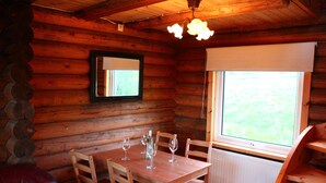 A wooden cabin interior with a dining table set for four, a mirror on the wall, and a window with a view of greenery.