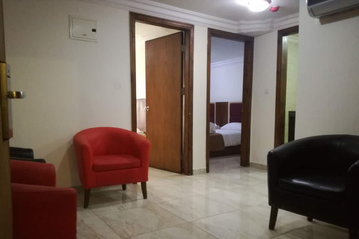 A hotel room with a bed, a red armchair, a black armchair, and a tiled floor.
