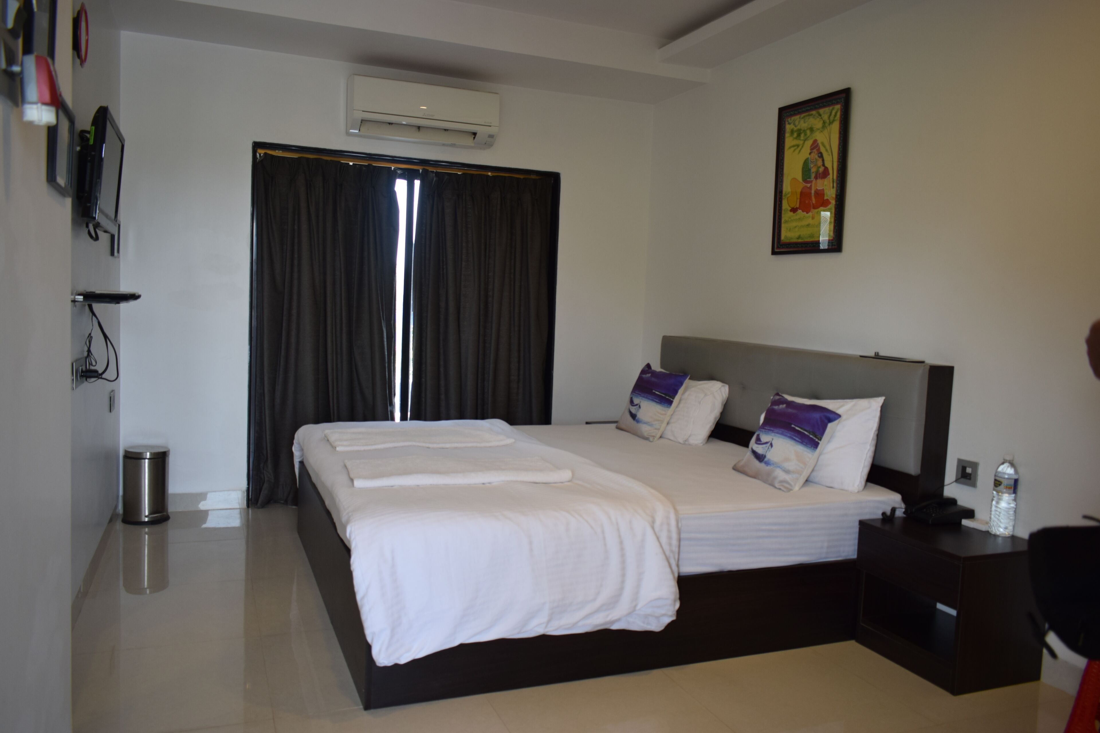 A hotel room with a large bed, a bedside table, a trash bin, and a wall-mounted air conditioner.