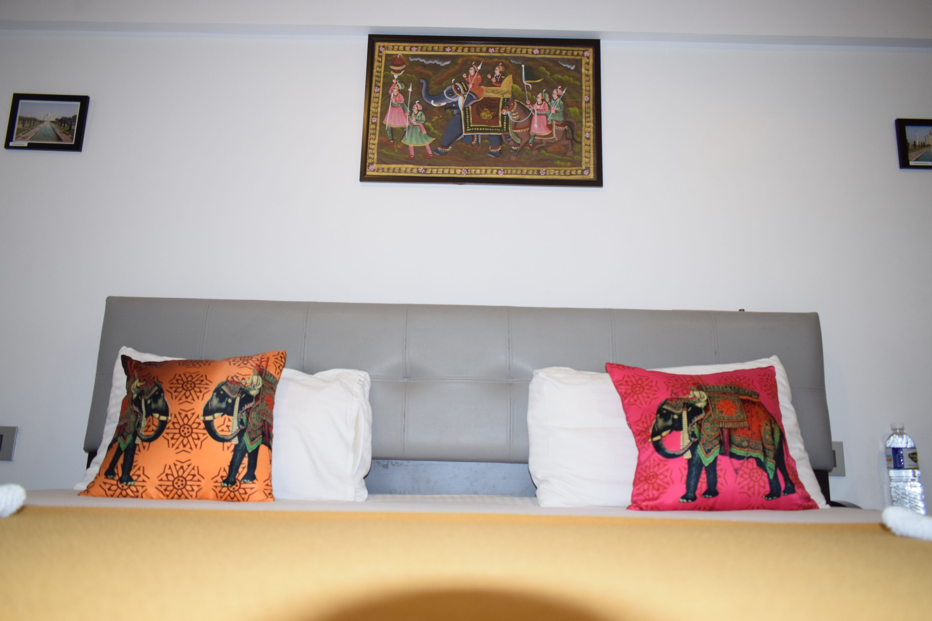 A hotel room with a bed featuring two decorative pillows, a framed artwork on the wall, and a water bottle on the nightstand.