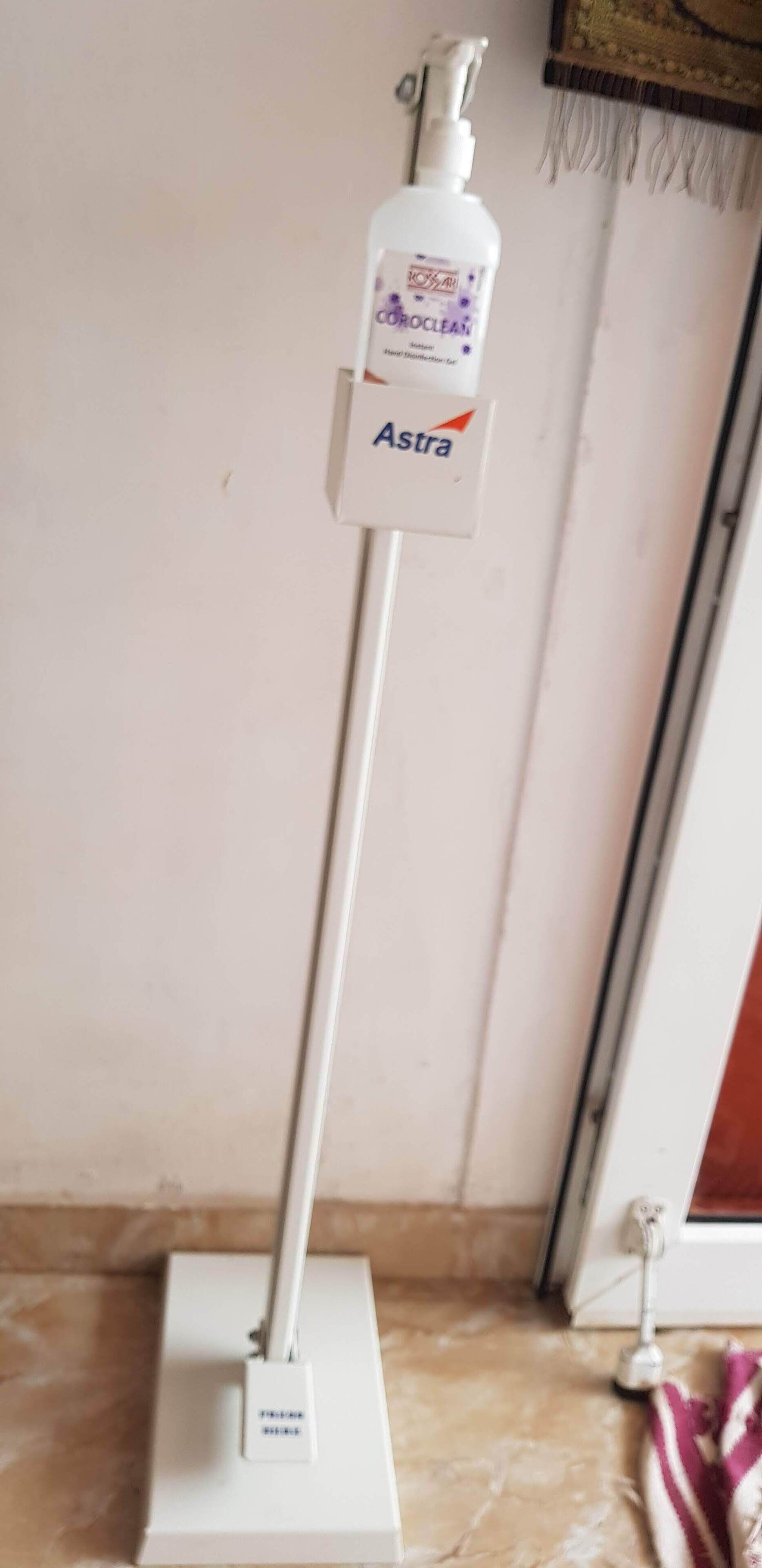A white floor lamp with a bottle of Astra hand sanitizer attached to it.