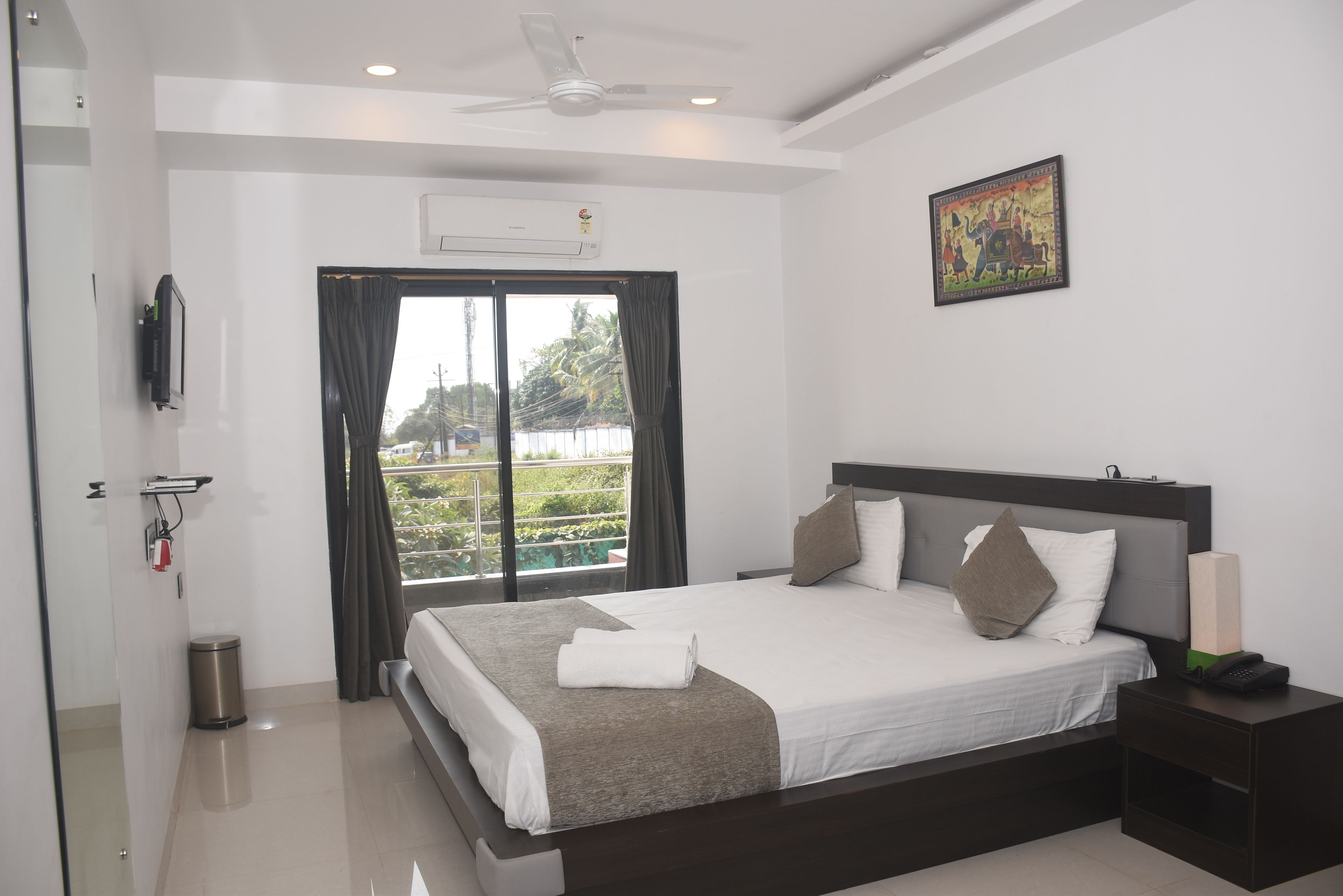 A modern hotel room with a large bed, a ceiling fan, an air conditioning unit, a small trash bin, and a view of greenery through the window.