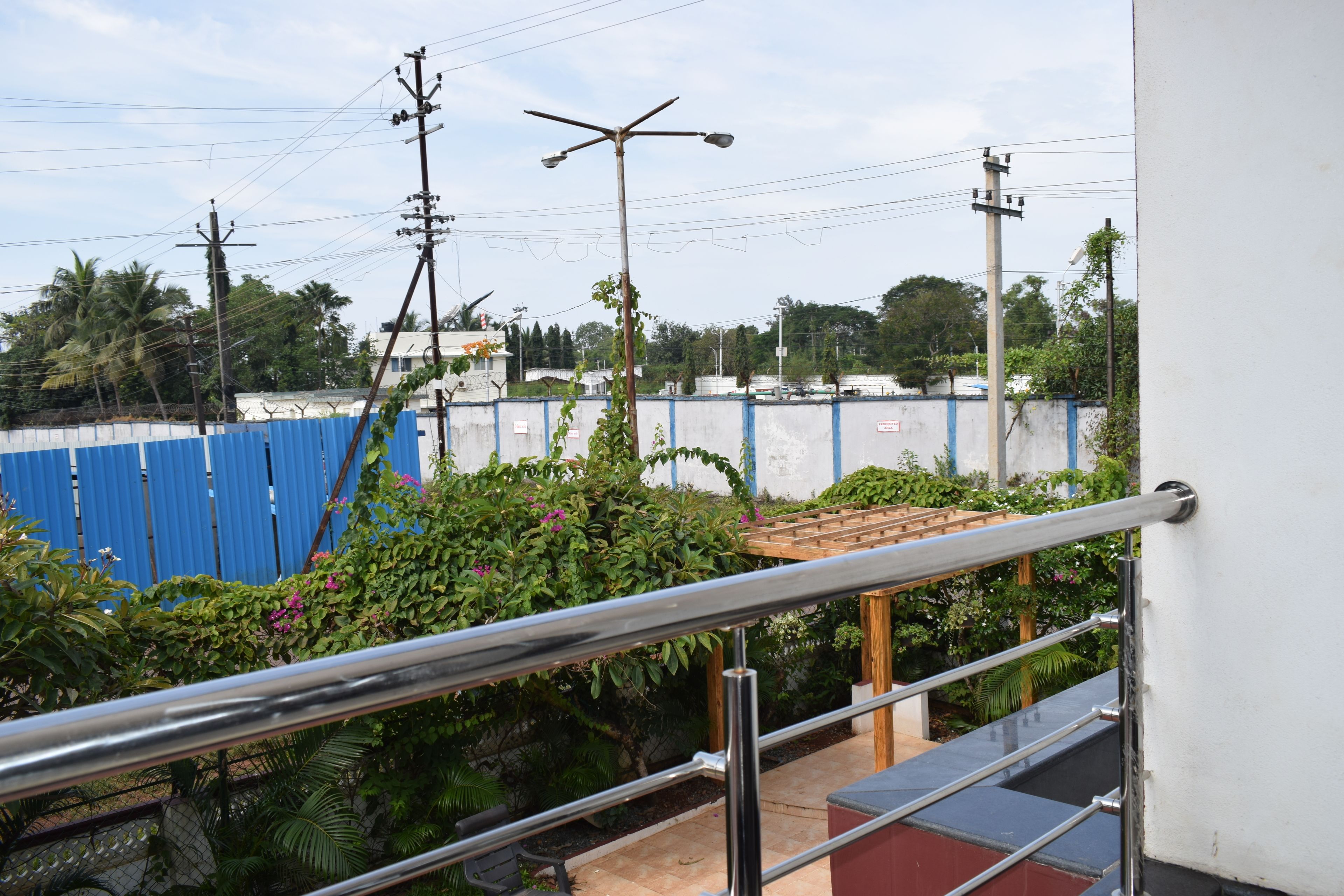 A view from a balcony with a metal railing, overlooking a blue fence, greenery, and a clear sky.