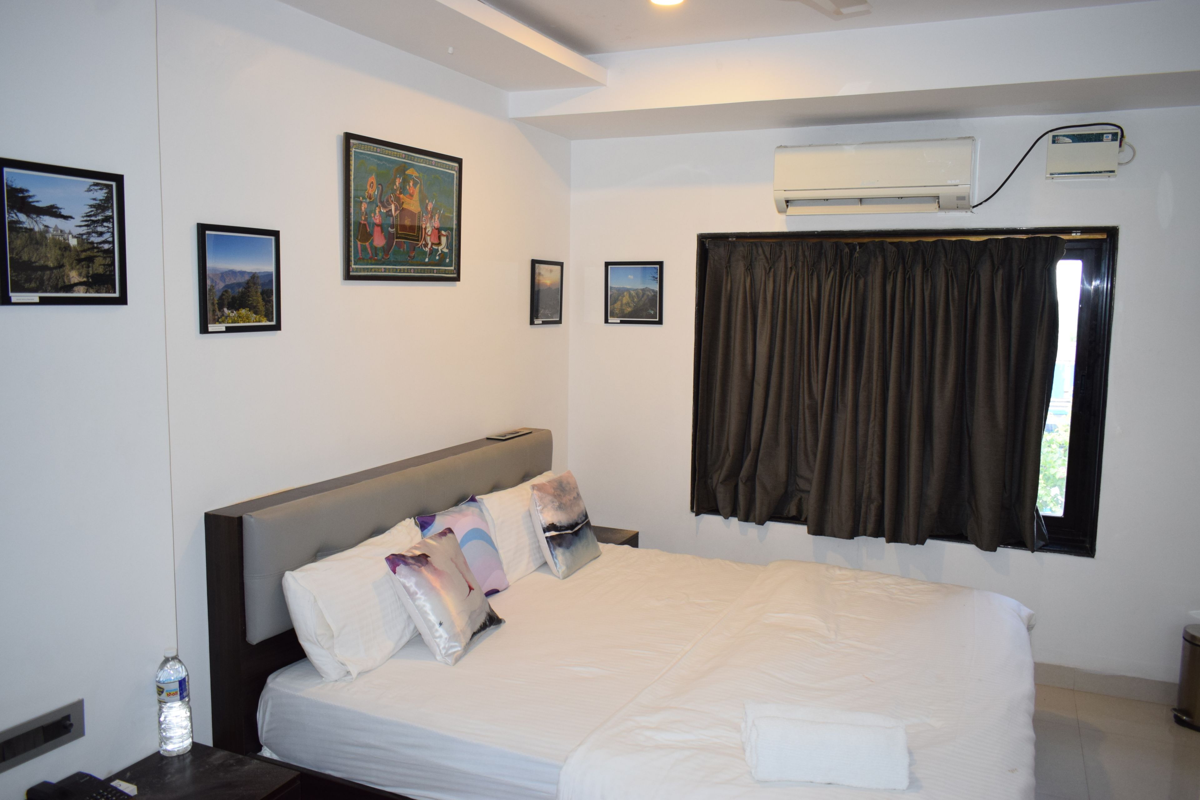 A hotel room with a large bed, a nightstand with a bottle of water, a remote control, and framed pictures on the wall.