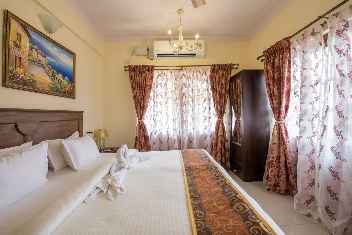 Habitación de hotel con cama, cabecera de madera, un cuadro en la pared, unidad de aire acondicionado y cortinas.