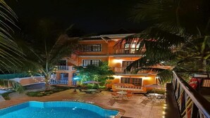 Une vue nocturne d’un hôtel doté d’une piscine, d’un espace extérieur aménagé avec des sièges et d’une terrasse bien éclairée.