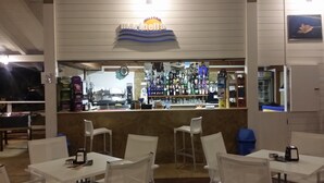 A bar area with a counter, various bottles, and a clock. There are white chairs and tables in the foreground.