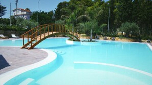 A swimming pool with a wooden bridge, surrounded by trees and a building in the background.