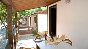 A covered outdoor dining area with wooden tables and chairs, a white wall, and a view of the surrounding trees.