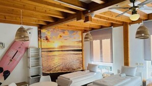 Une pièce chaleureuse avec un plafond en bois, un grand tableau représentant un coucher de soleil, un canapé blanc et une table à manger avec des chaises.