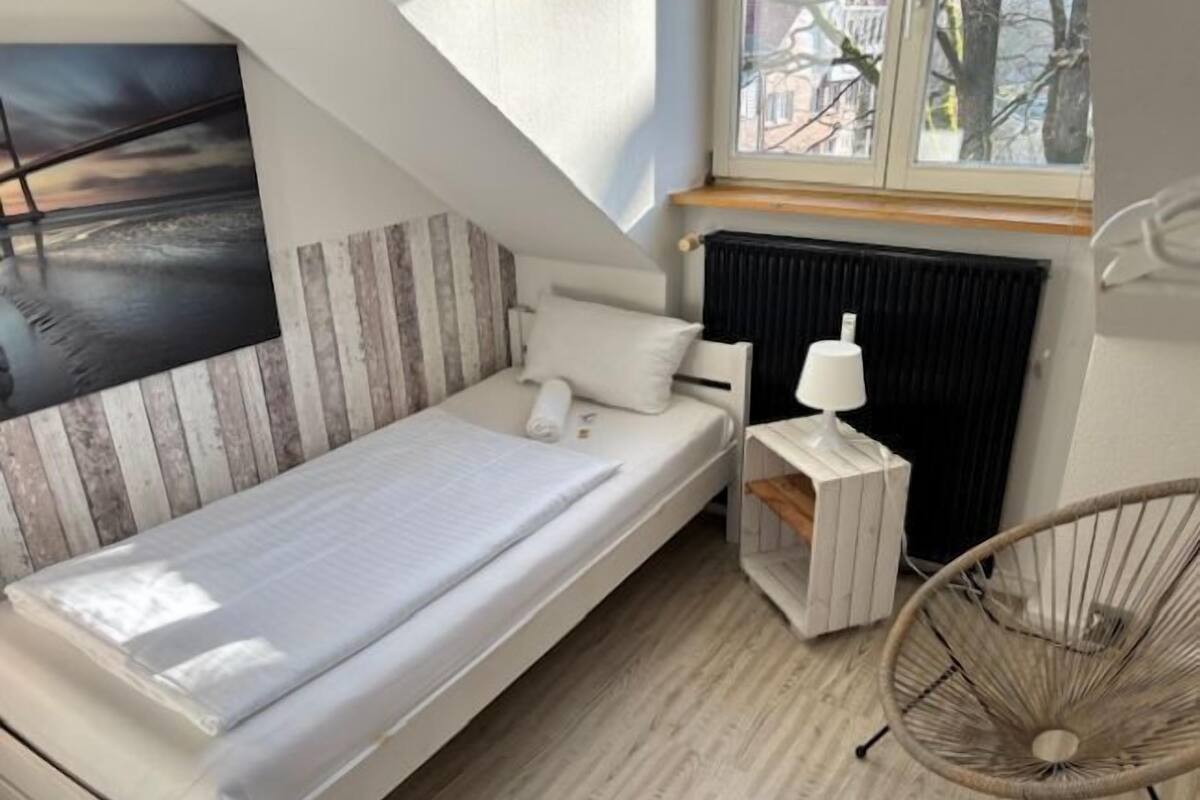 A small, modern bedroom with a single bed, a window with a view of trees, a radiator, and a chair.