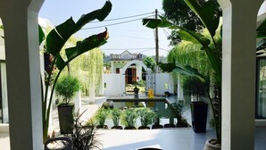 Une vue depuis un balcon orné de plantes en pot, avec un sol carrelé et un bâtiment à l’arrière-plan, caractérisé par une façade blanche et une arche.