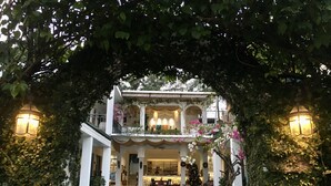 Une maison à deux étages dotée d’un balcon, entourée de verdure et éclairée par des lampes fixées aux murs.