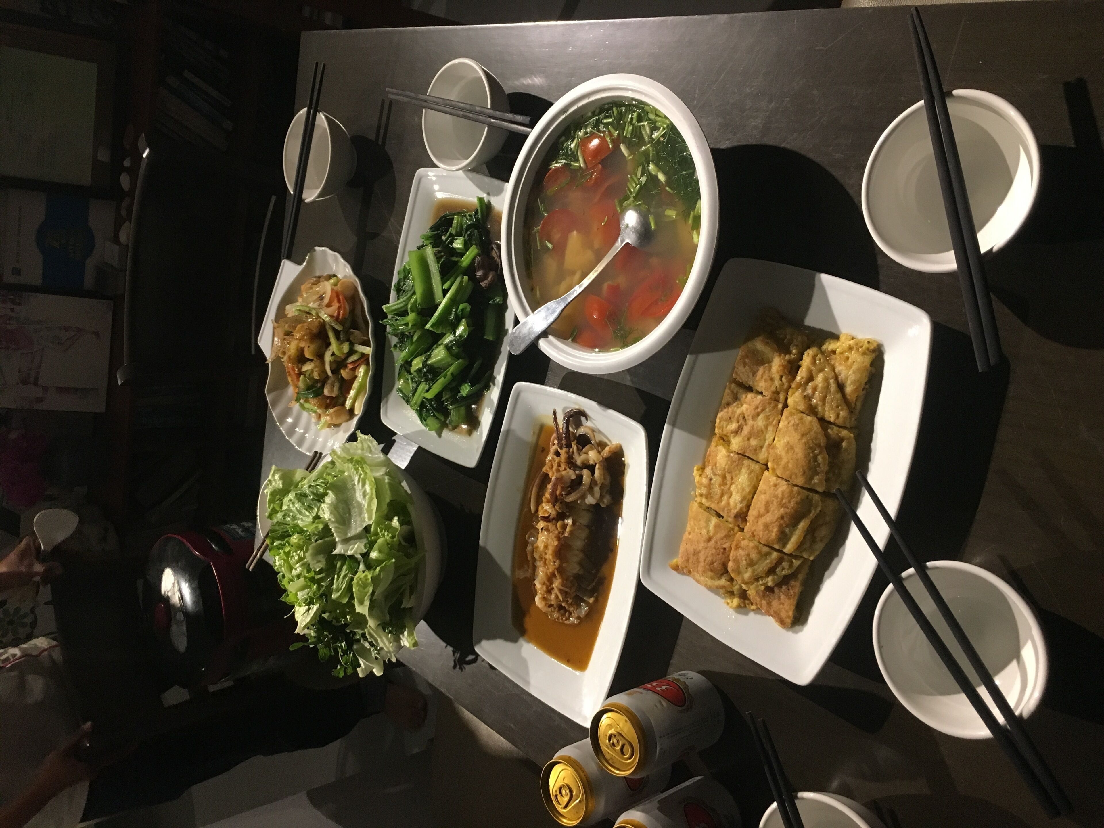 Una mesa con varios platos, incluyendo sopa, verduras y carne, junto con palillos y una lata de bebida.
