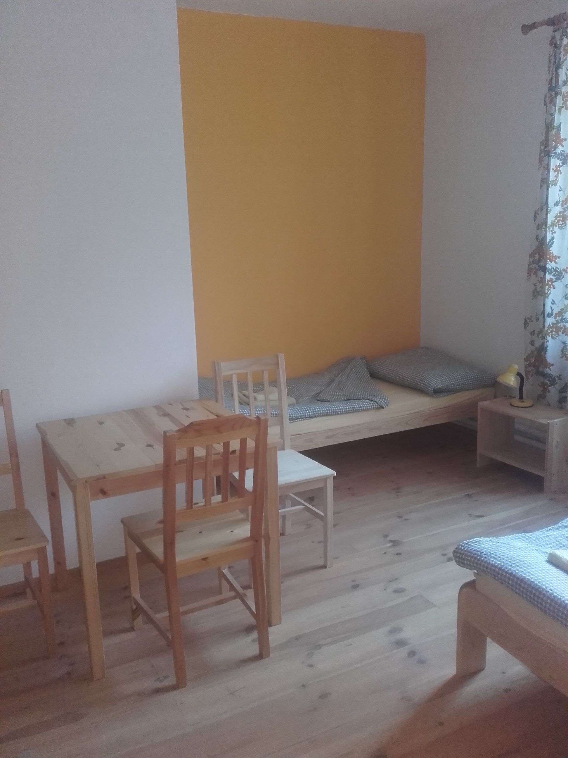 A room with a wooden floor, a bed with a checkered bedspread, a small table with chairs, and a wall with a yellow section.