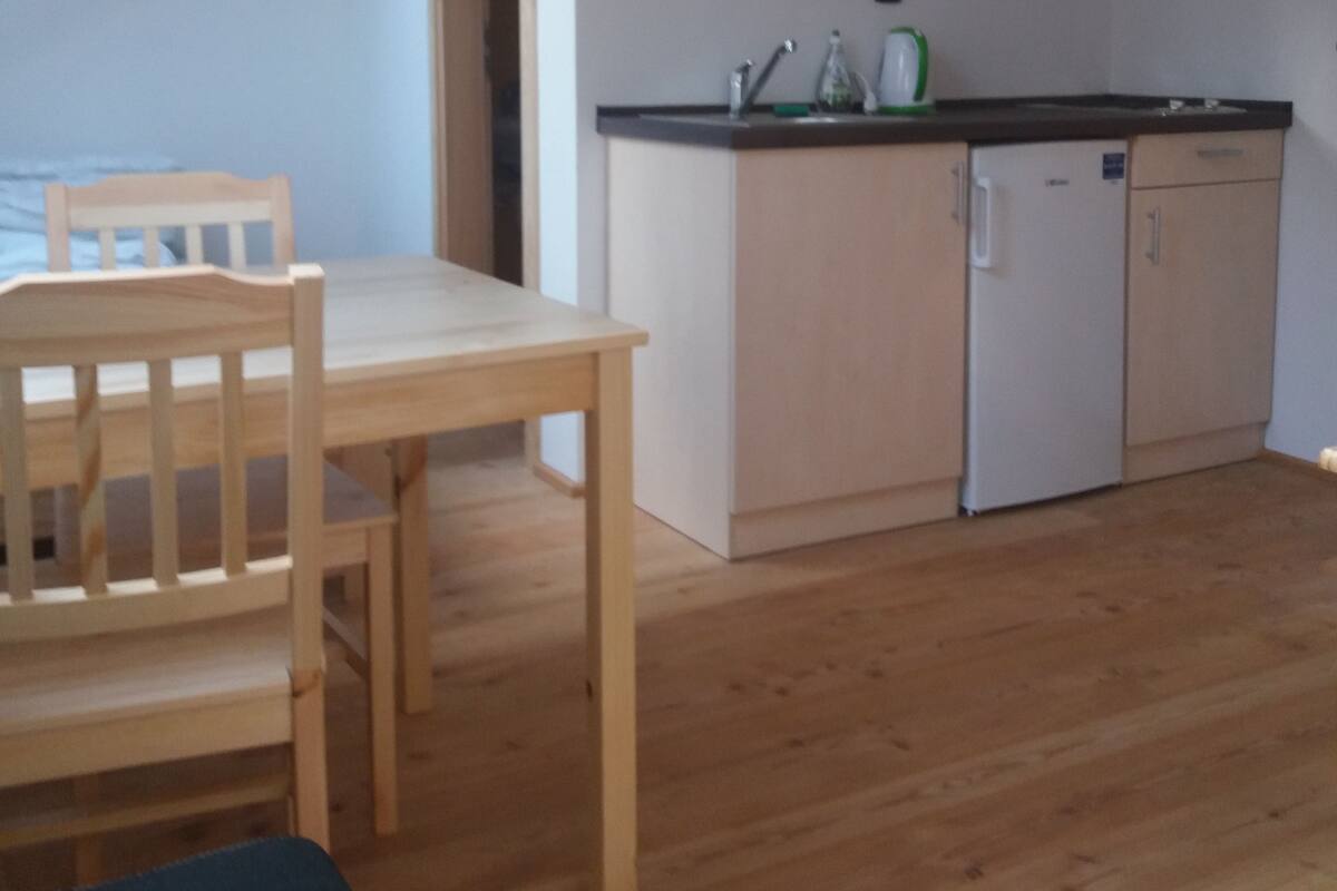 A compact kitchen with wooden cabinets, a sink, and a dishwasher. There is a dining table with chairs and a bench.