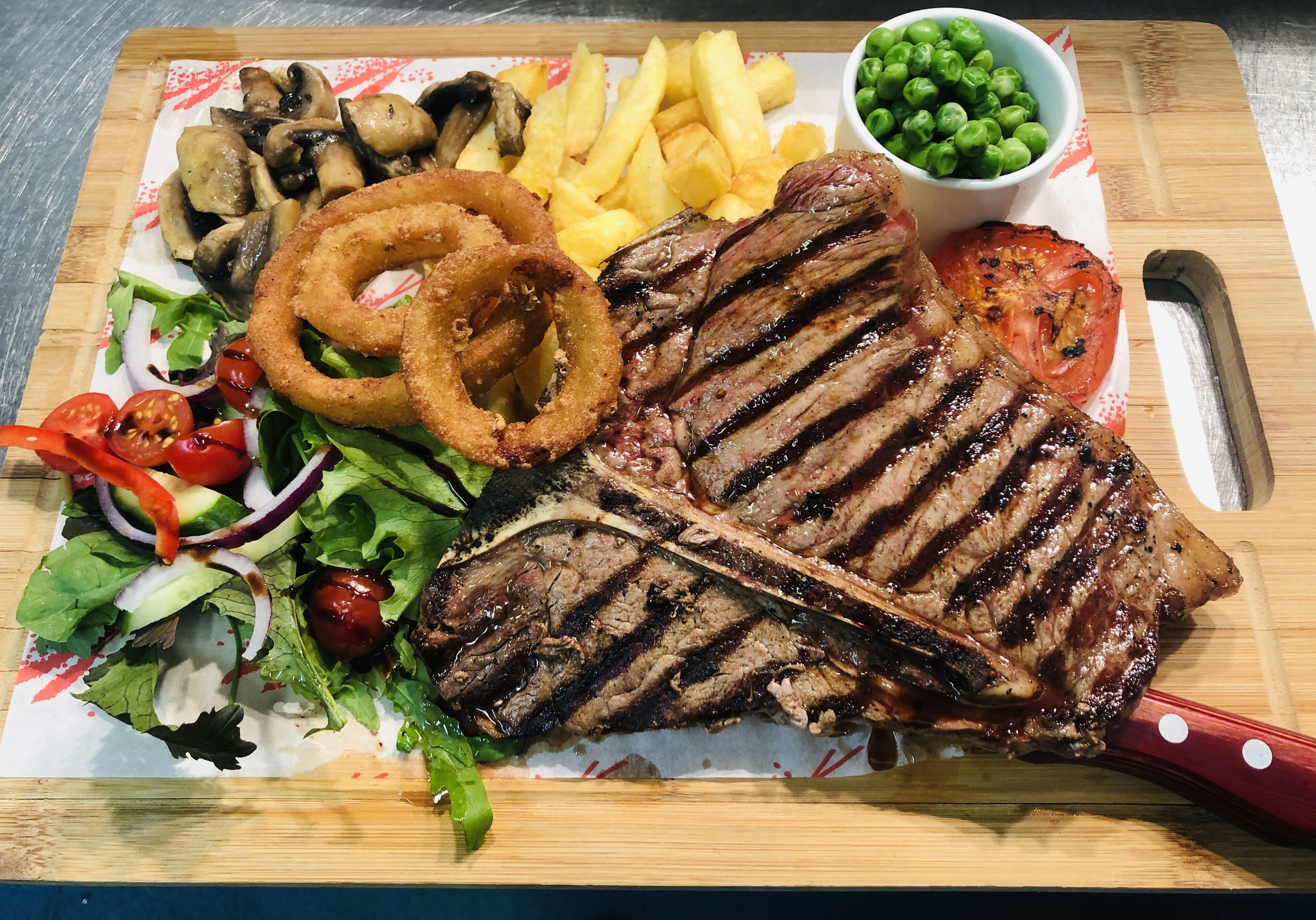 Un bistec con marcas de parrilla, aros de cebolla, papas fritas, ensalada y un tomate sobre una tabla de madera.