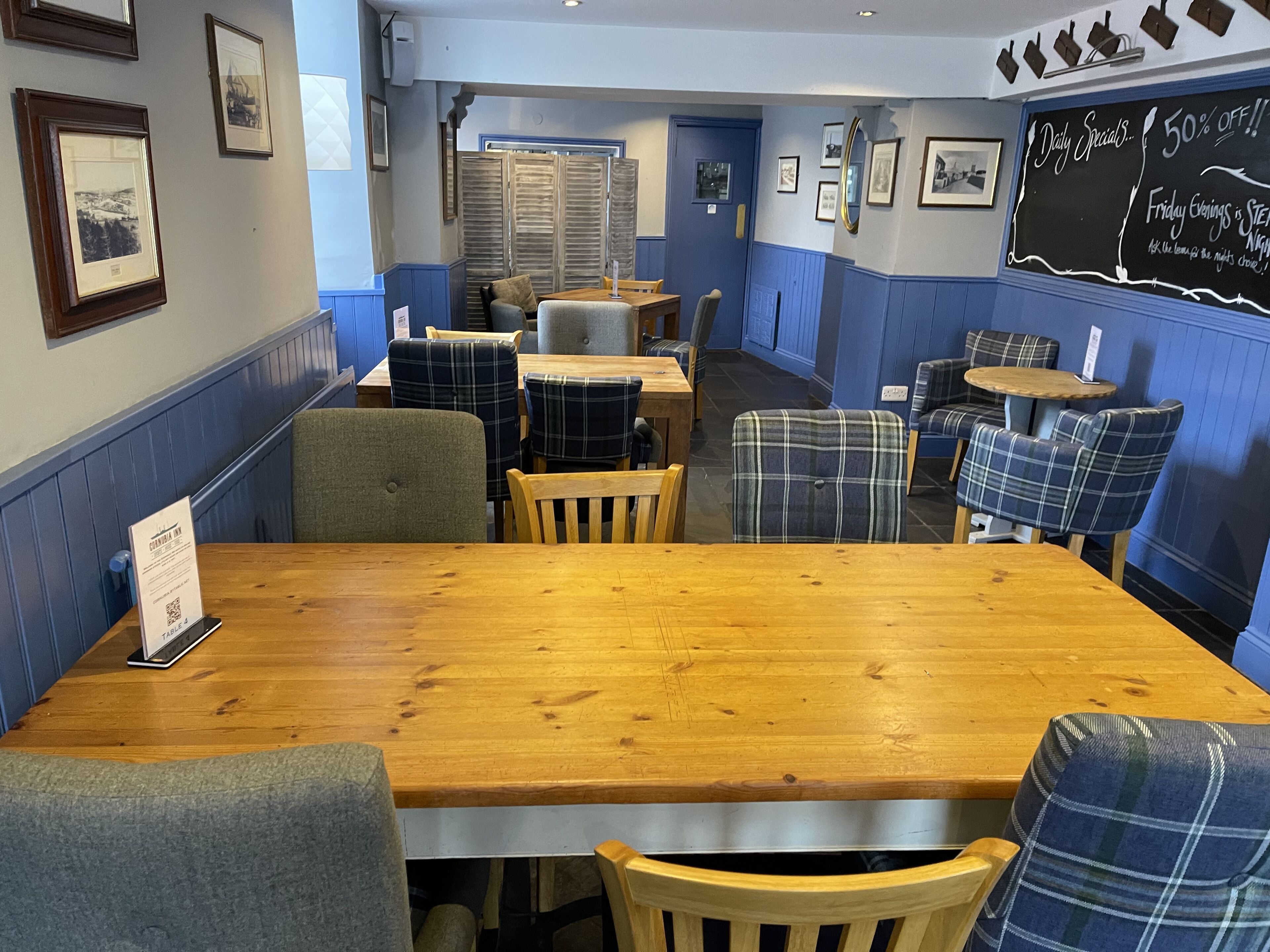 A cozy restaurant interior with wooden tables and plaid chairs, framed pictures on the walls, and a chalkboard with specials.