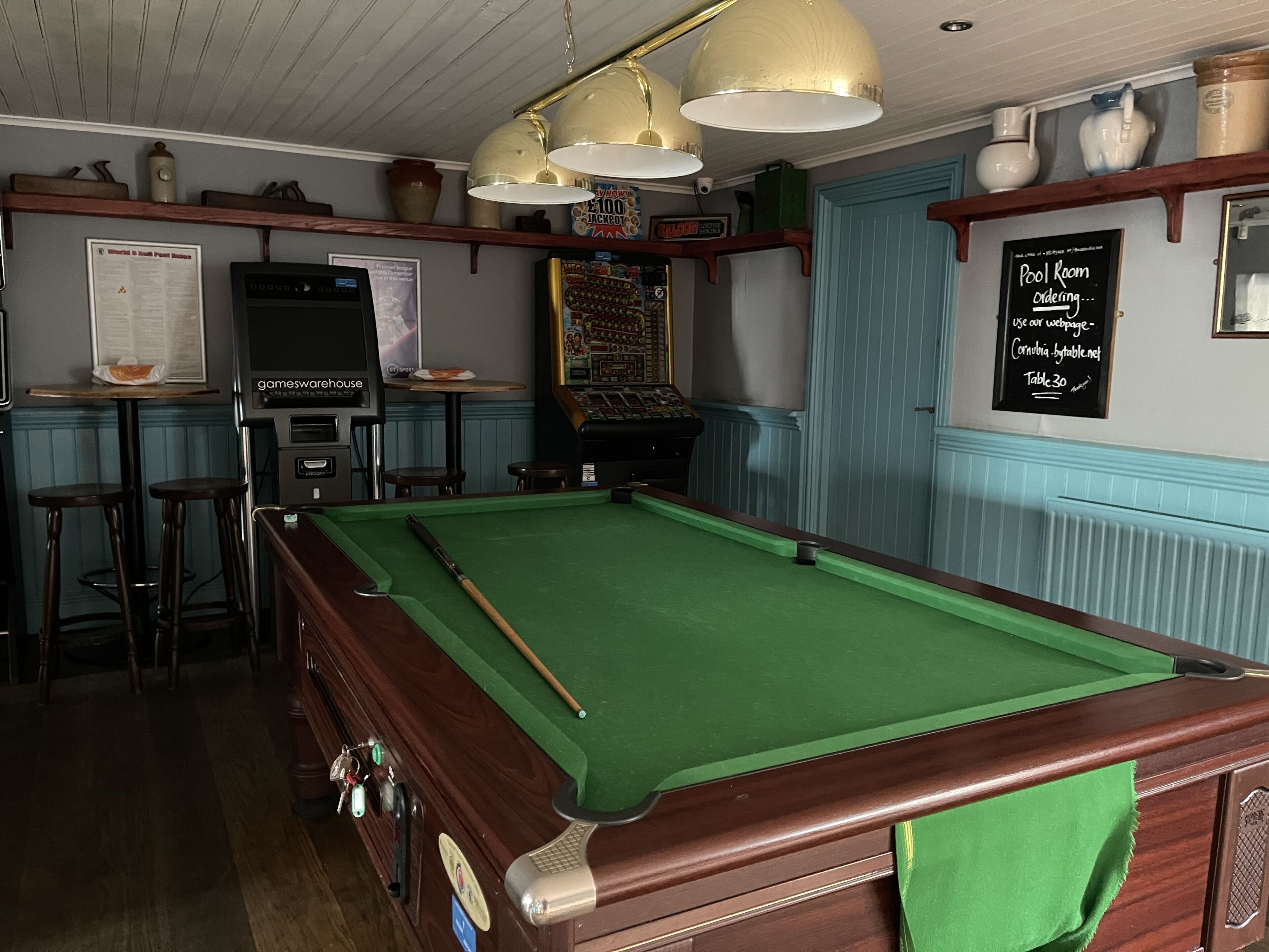 Une table de billard recouverte d’un drap vert, avec un cadre en bois et des luminaires suspendus au plafond.