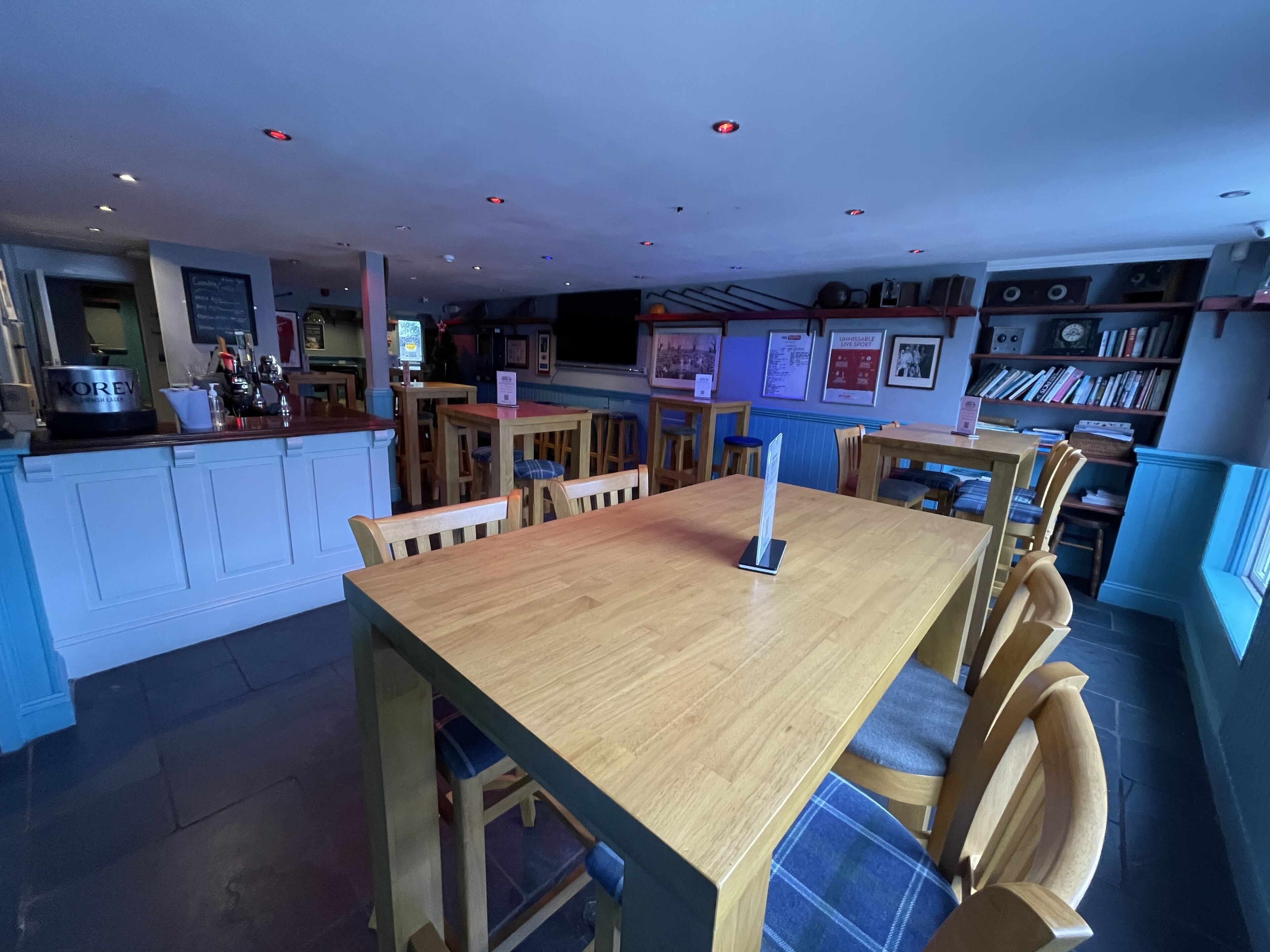 A restaurant with wooden tables and chairs, a bar area, and a bookshelf with books.