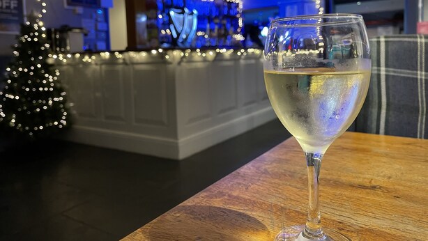 Ein Glas Weißwein auf einem Holztisch, im Hintergrund ein Weihnachtsbaum und eine Bar.