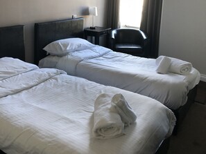 A hotel room with two beds, each with white linens and towels. There is a small table with a lamp, a chair, and a wall-mounted light fixture.