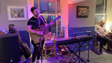 Ein Musiker spielt Gitarre und Keyboard in einem Raum mit Fenster, Stuhl und einem kleinen Weihnachtsbaum.
