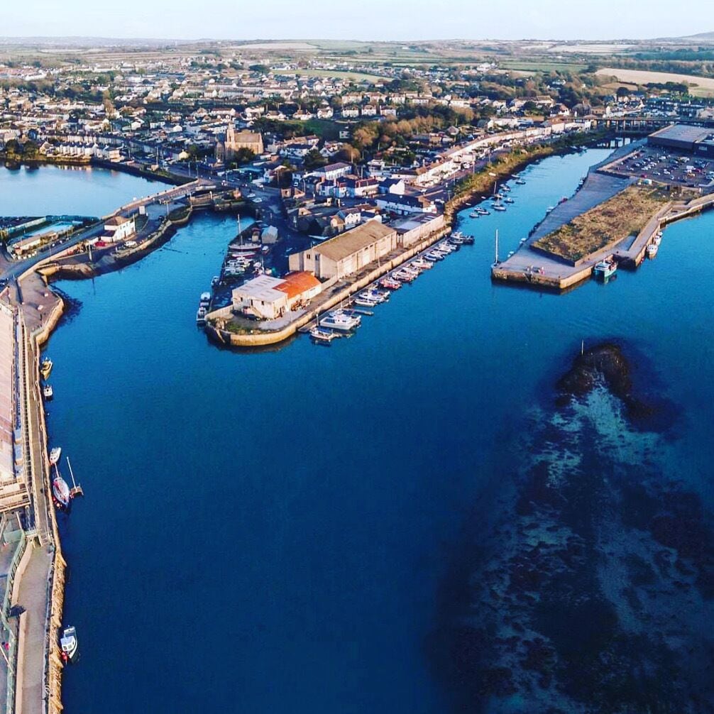 An aerial view of a coastal town with a marina, a large building with a red roof, and a bridge connecting to the mainland.