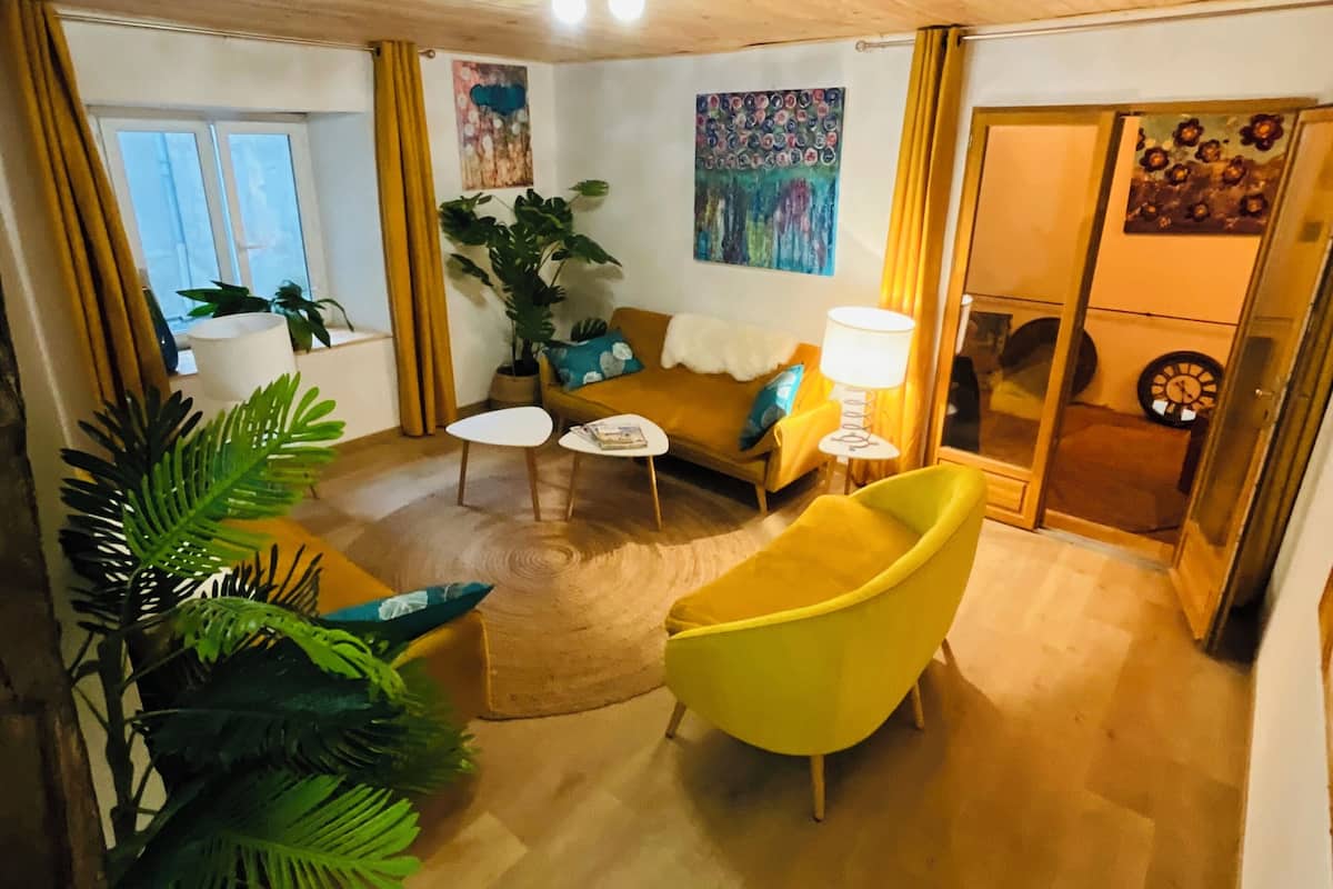A cozy living room with wooden flooring, a yellow armchair, a sofa, and a coffee table. There are two paintings on the wall, a potted plant, and a door leading to another room.