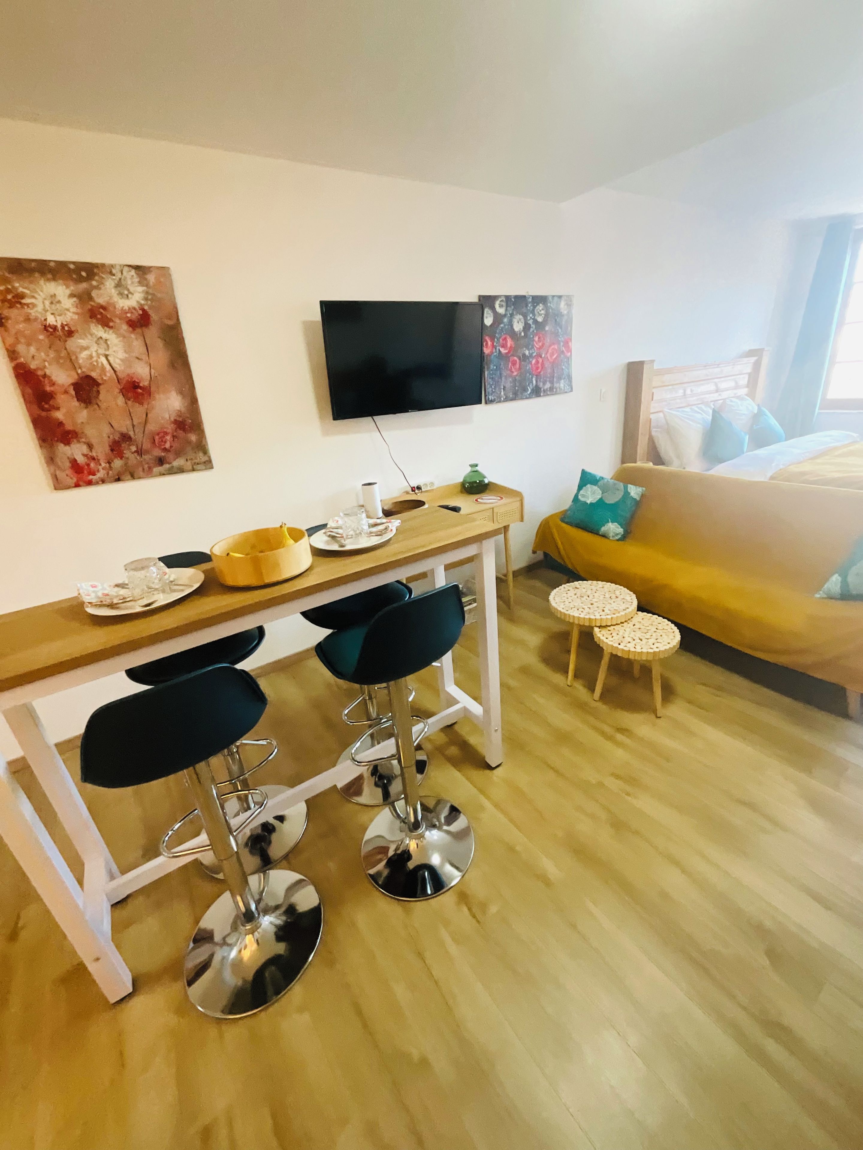 A modern living room with a wooden dining table, two bar stools, a yellow sofa, a TV mounted on the wall, and a small round table.