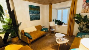 A cozy living room with a wooden ceiling, a sofa, a yellow armchair, a small round table, and a window with curtains. - Labyrinthe (Mirepoix)