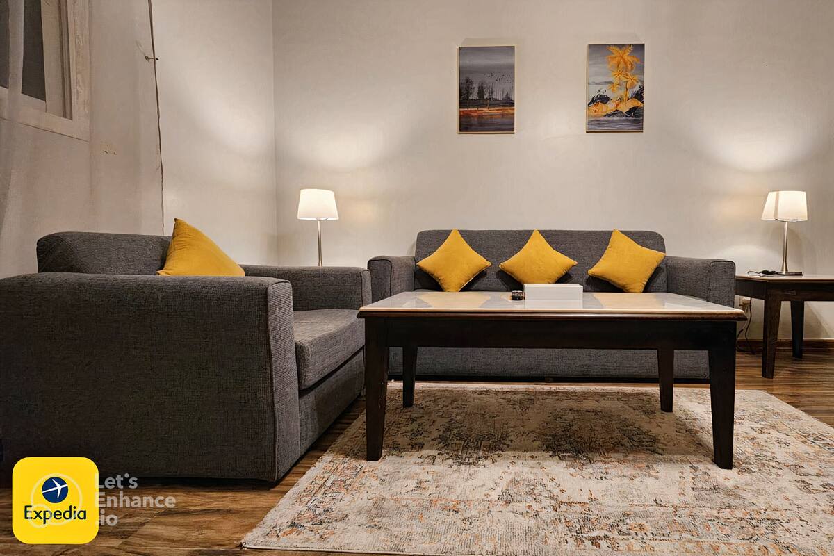 A living room with a grey sofa and armchairs, a coffee table, two lamps, and a rug. There are two paintings on the wall.